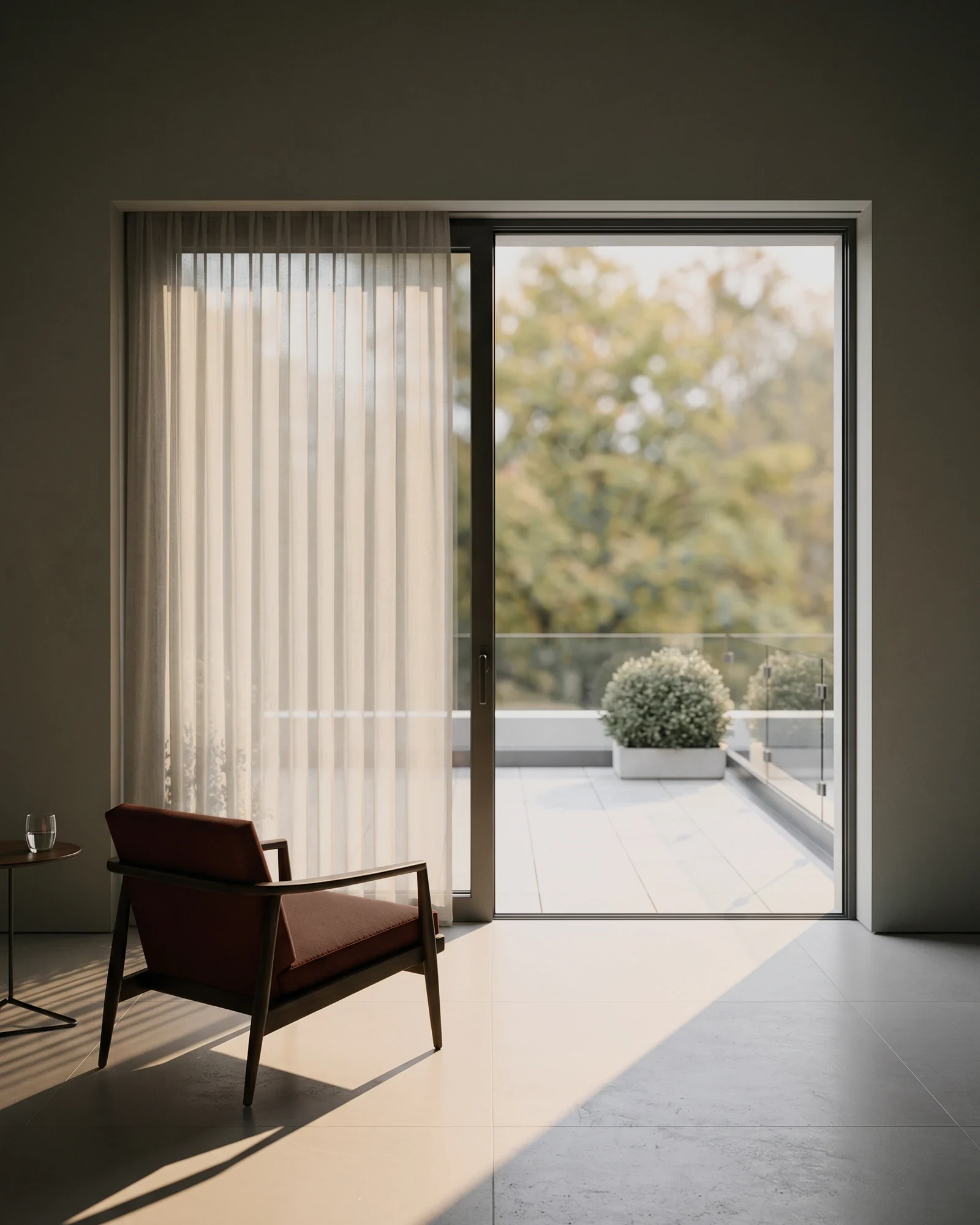 A room in shadow with its terrace door slid open. An armchair faces the light, and a glass stands on the side table beside it.