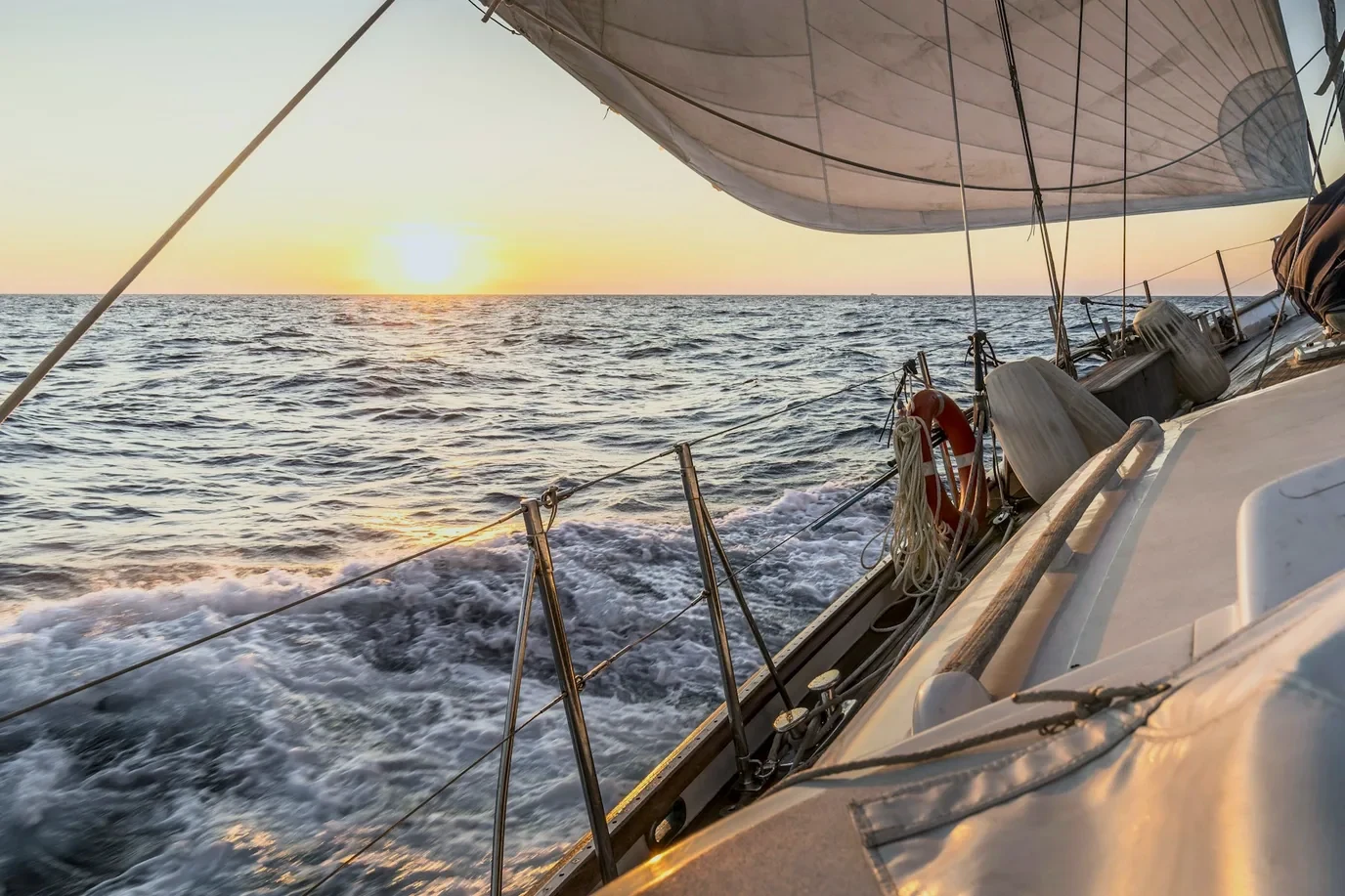 The deck of a sailing boat under way. The sail is up, the sun sits low over the water and the wake runs out behind.