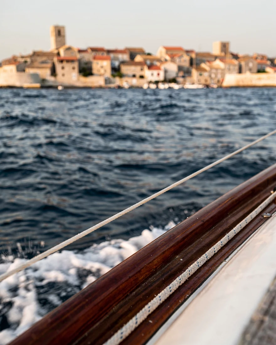 An old coastal town seen across the water from a moving boat. The varnished rail and the wake fill the foreground.